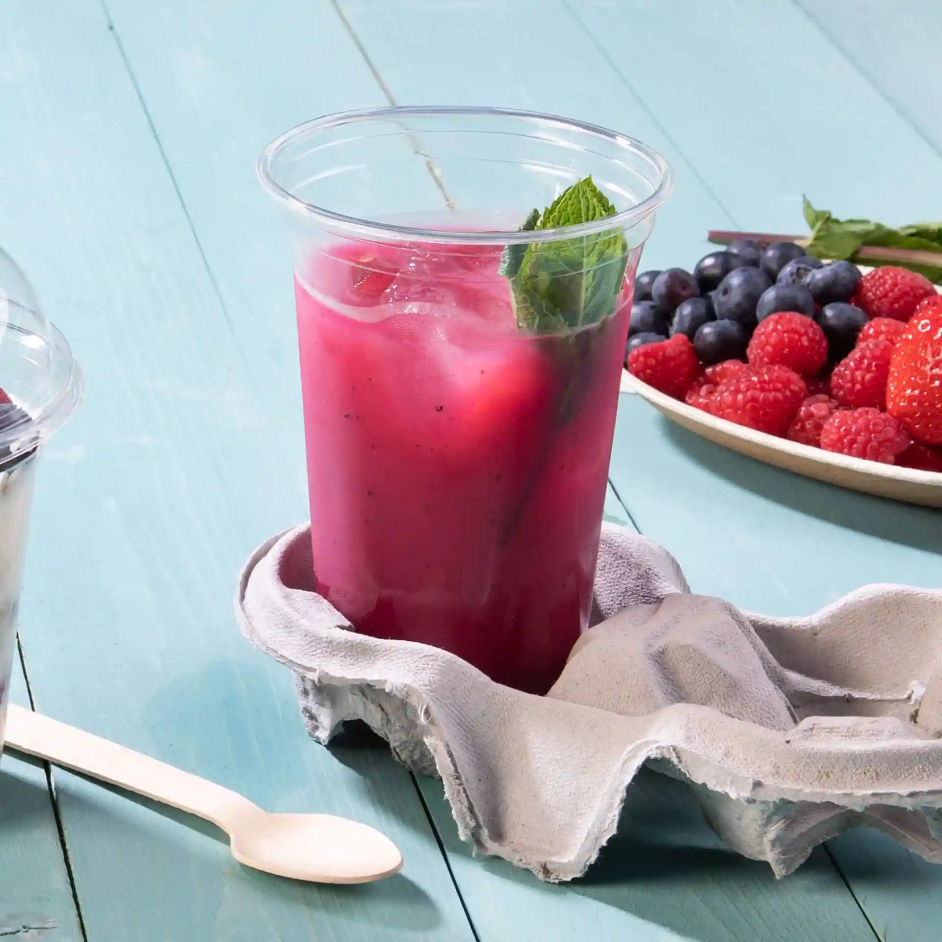 Clear rPET cup filled with a pink beverage, garnished with mint, on a grey cloth with berries in the background.