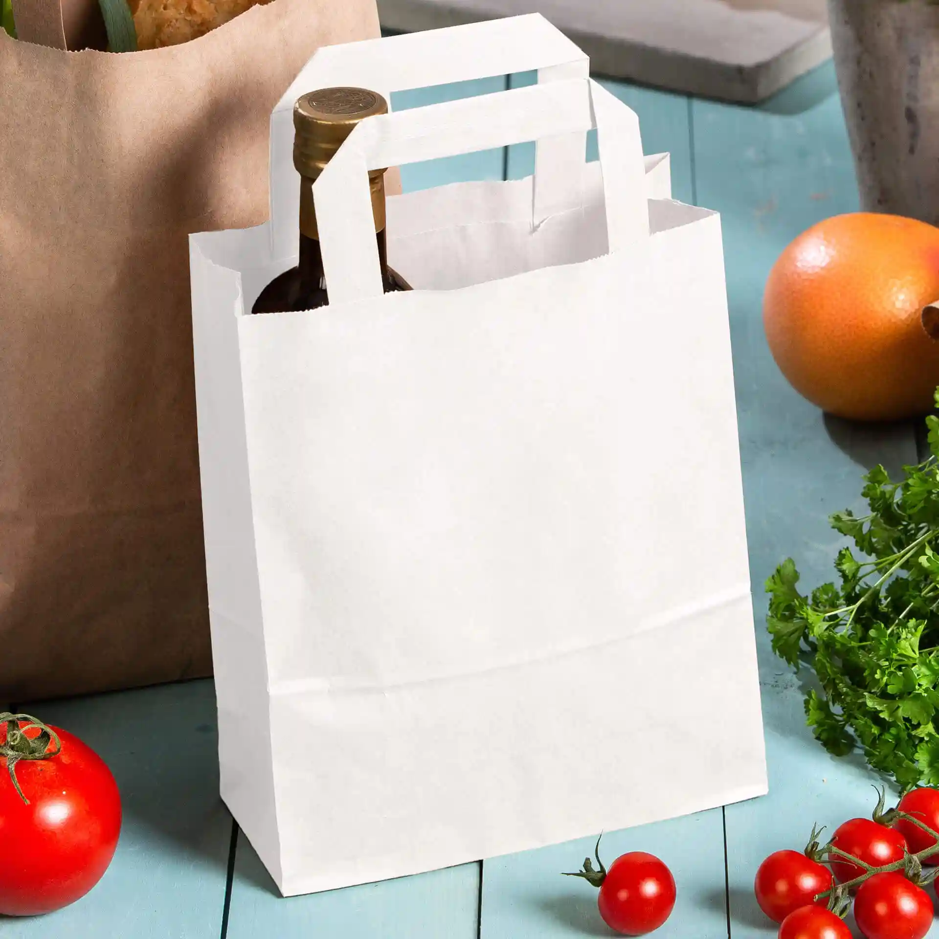 Weißer Kraftpapierbeutel mit Griffen, der eine Flasche hält, umgeben von Tomaten und einer Orange auf einer blauen Fläche.