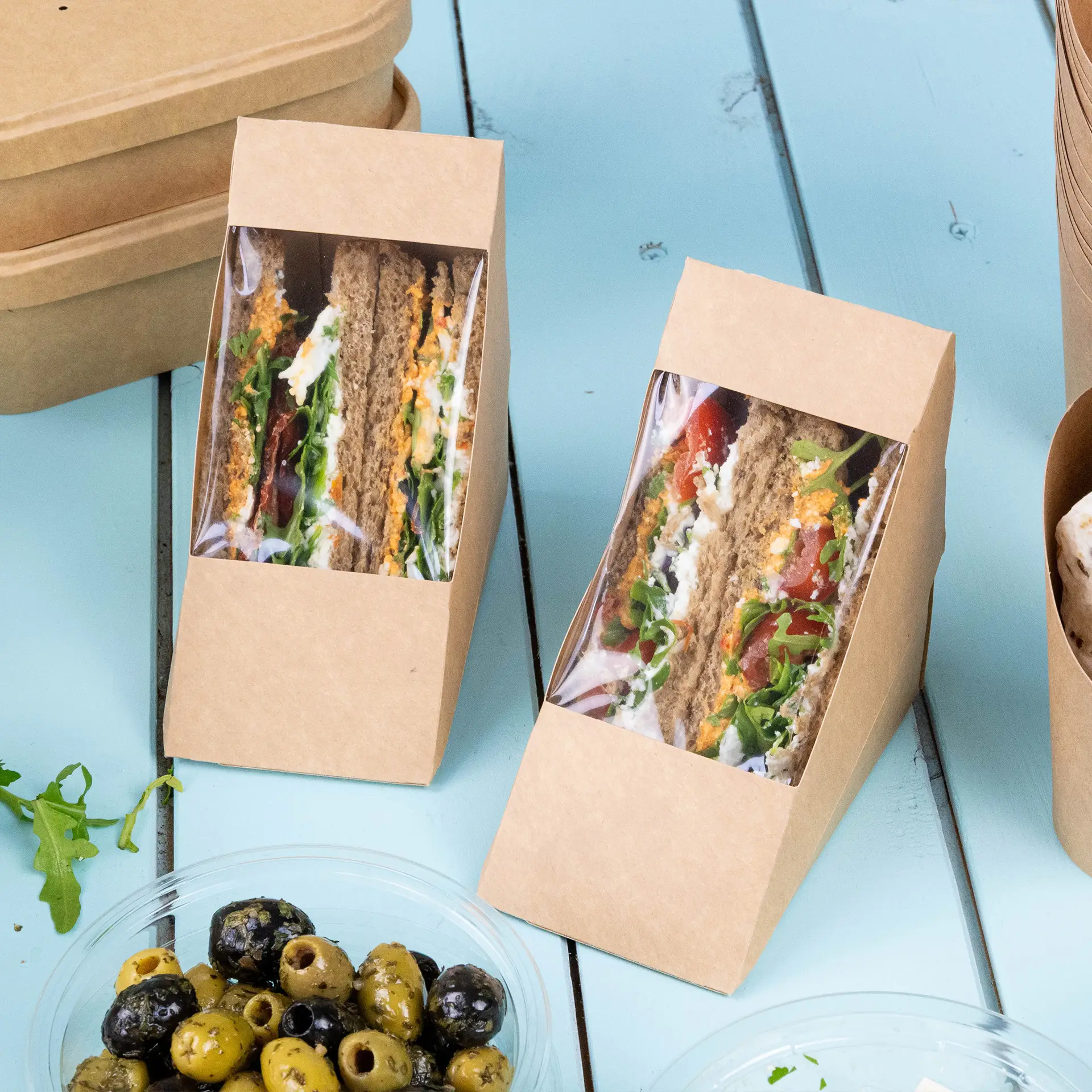 Zwei braune Papp-Sandwich-Winkel mit transparentem PLA-Fenster, das Sandwiches und gefüllte Brötchen auf einer blauen Fläche zeigt, mit Oliven in einer Schüssel daneben.
