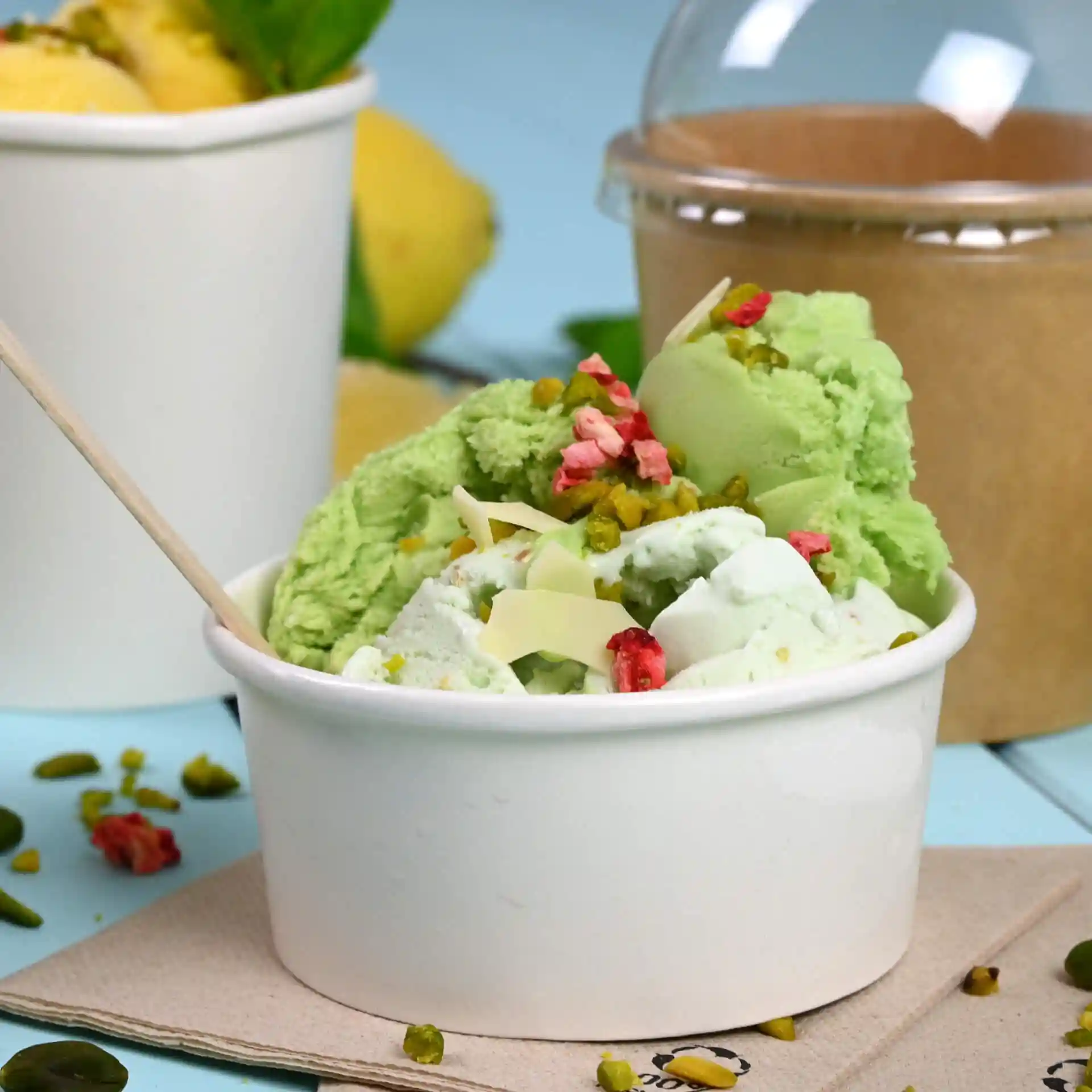 White paper ice cream cup filled with green and white ice cream, topped with nuts and pink pieces, with a wooden spoon.
