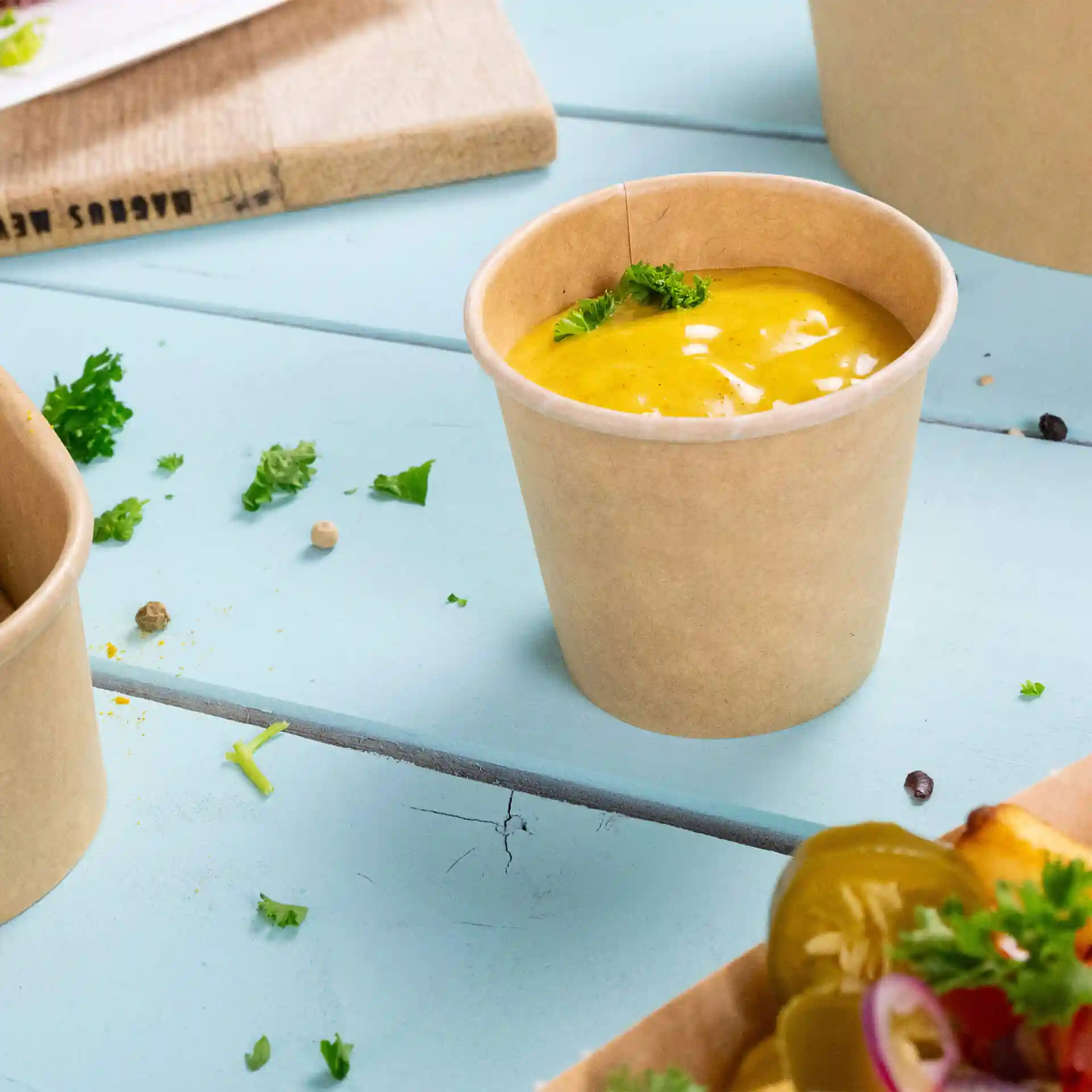 Brown round deli container with a lid, filled with yellow sauce and garnished with parsley, on a blue wooden surface.
