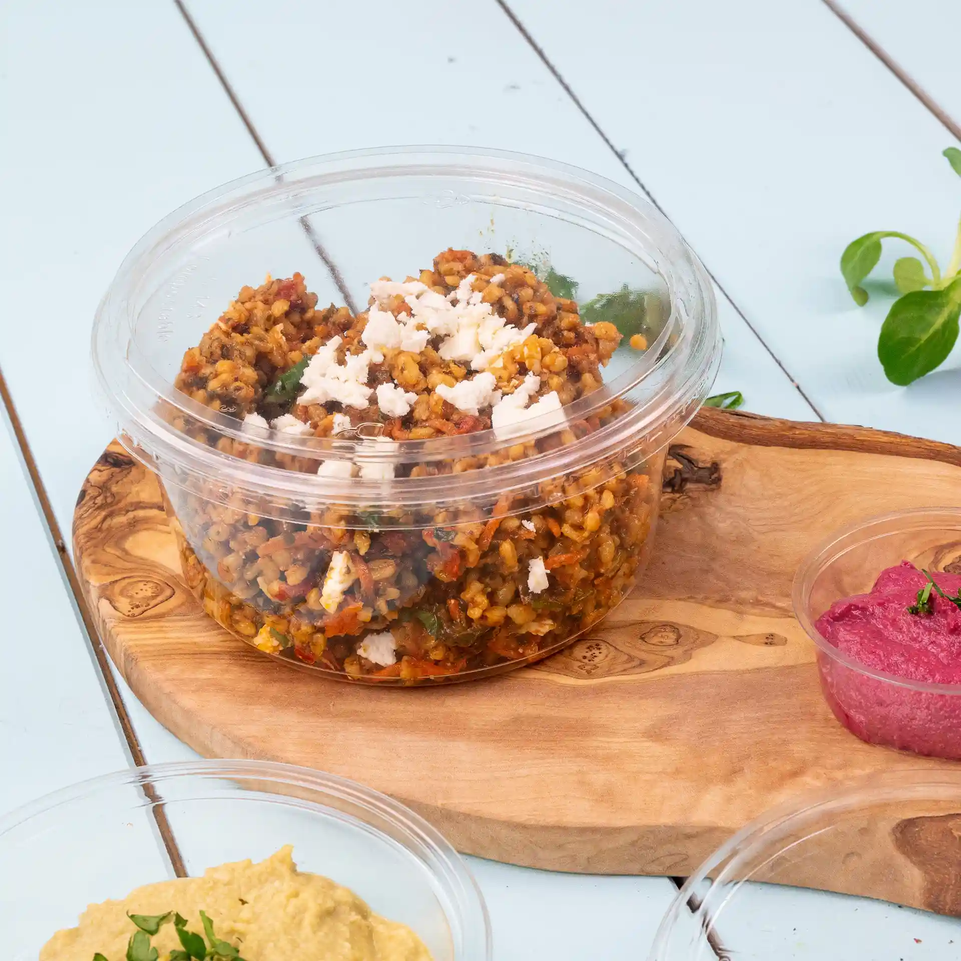 Transparent round deli cup with a lid containing a grain dish topped with feta cheese, placed on a wooden board.