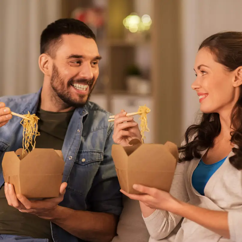 Paar genießt Takeout-Nudeln in braunen Boxen, mit Essstäbchen, zusammen in einer gemütlichen Innenumgebung.