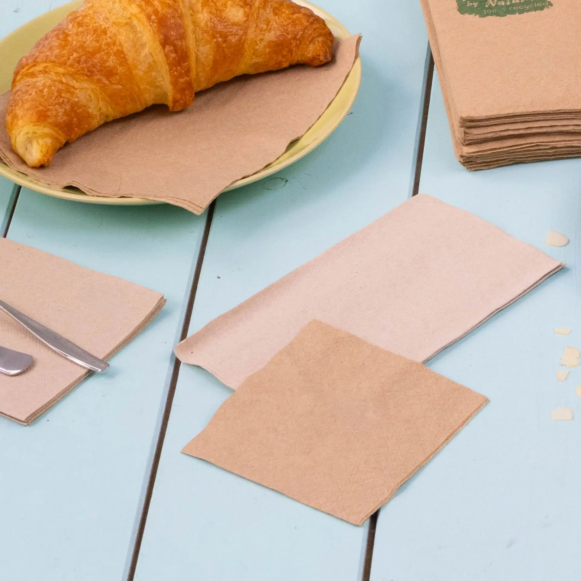 Bild eines Croissants auf einem gelben Teller mit mehreren braunen, recycelten Cocktailservietten auf einem blauen Tisch.