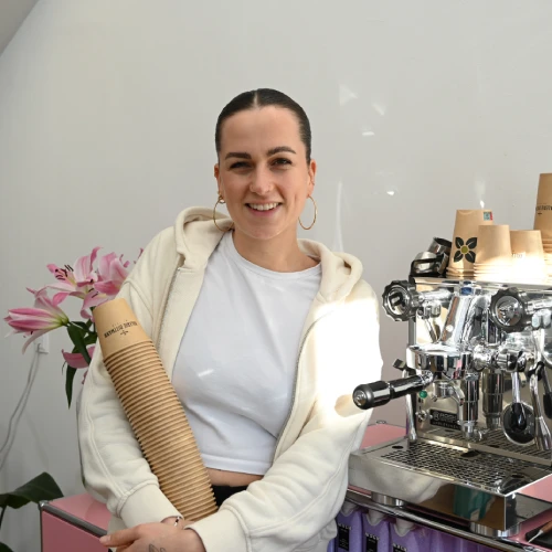 Frau in einem weißen Pullover, die Pappbecher hält, neben einer Espressomaschine und rosa Blumen im Hintergrund.