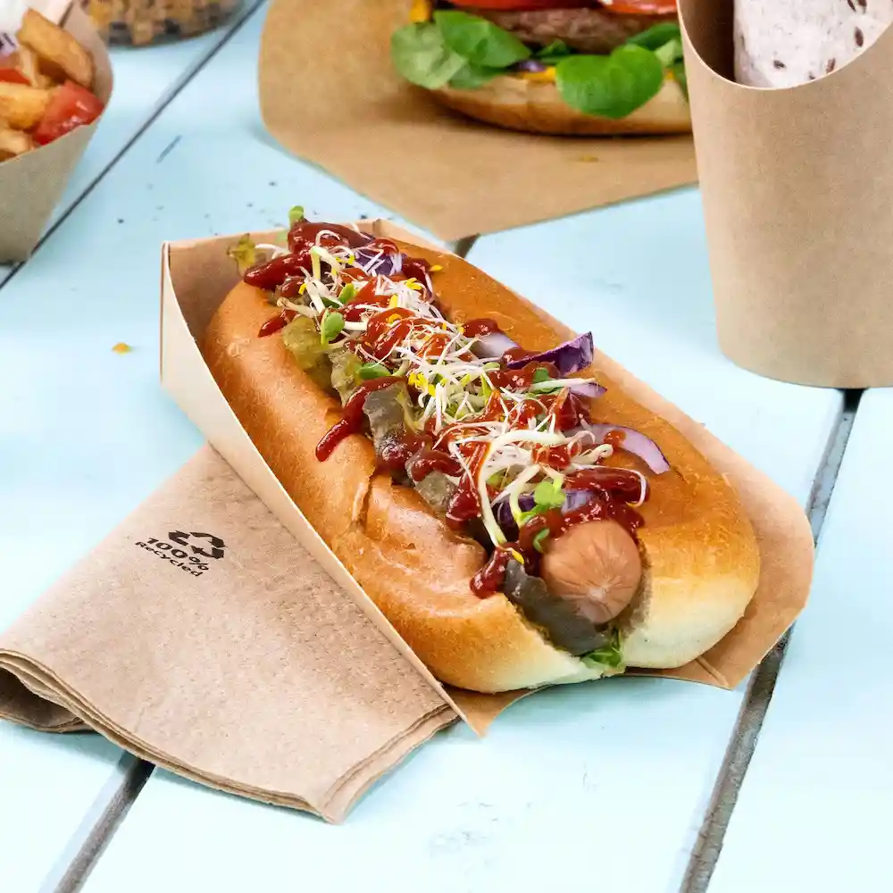 Hotdog in einem braunen Papierhalter, belegt mit Ketchup, Senf, Gurken und Sprossen, auf einem Holztisch.