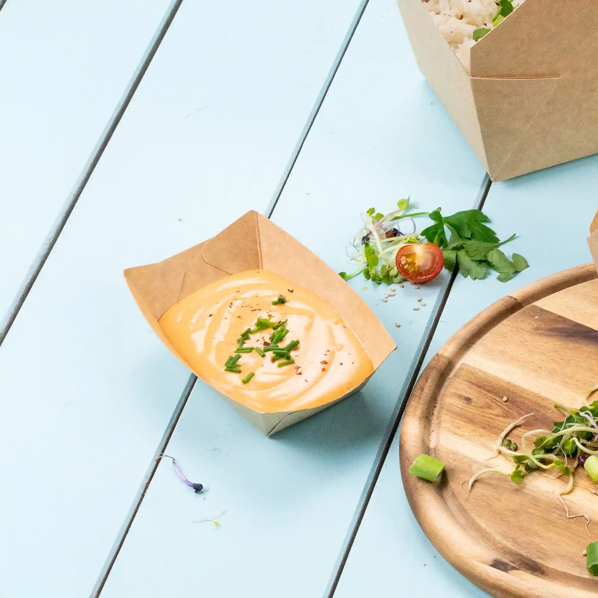 Brauner Papp-Snacktray mit einer cremigen Sauce, garniert mit gehacktem Schnittlauch, auf einer hellblauen Oberfläche.