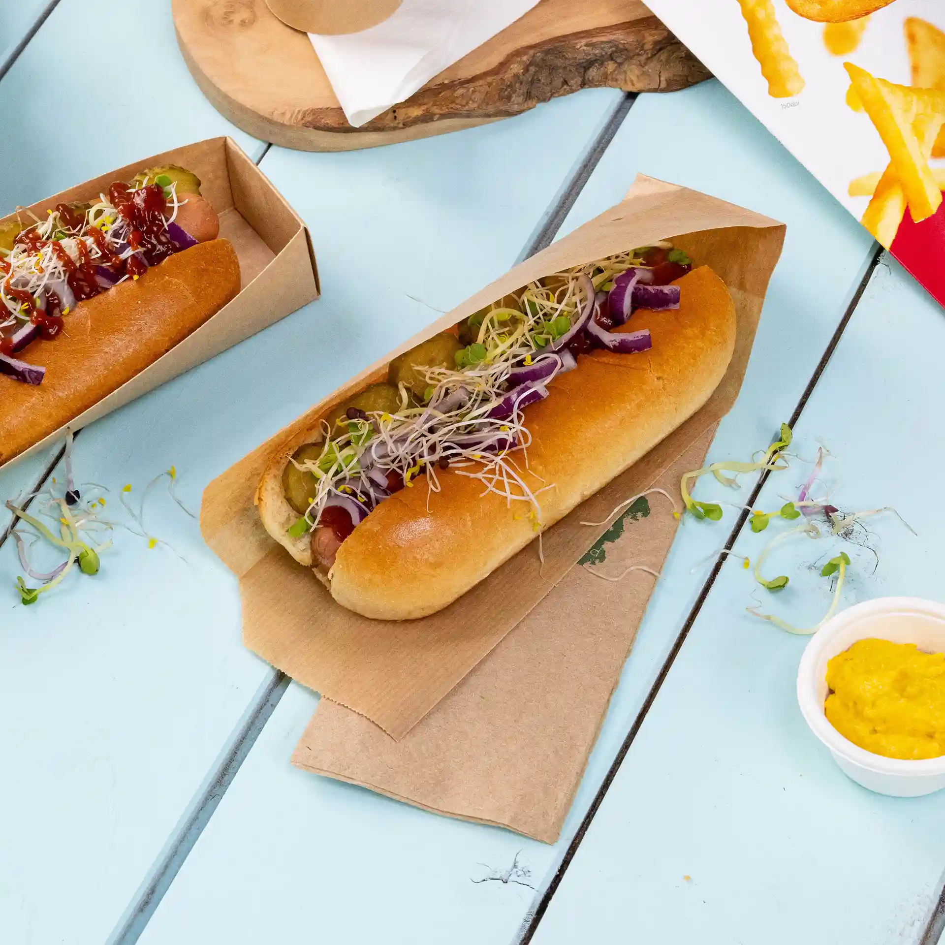 Zwei Hotdogs in braunen Pappschalen, belegt mit verschiedenen Gemüse und Saucen, auf einer hellblauen Oberfläche.