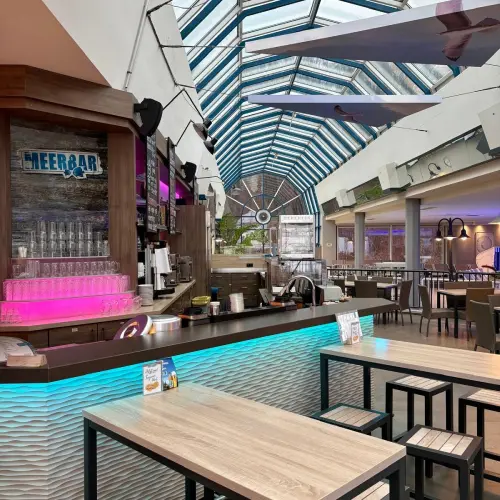 Interior view of a bar area with a glass roof, featuring a counter with glowing blue light and several tables.