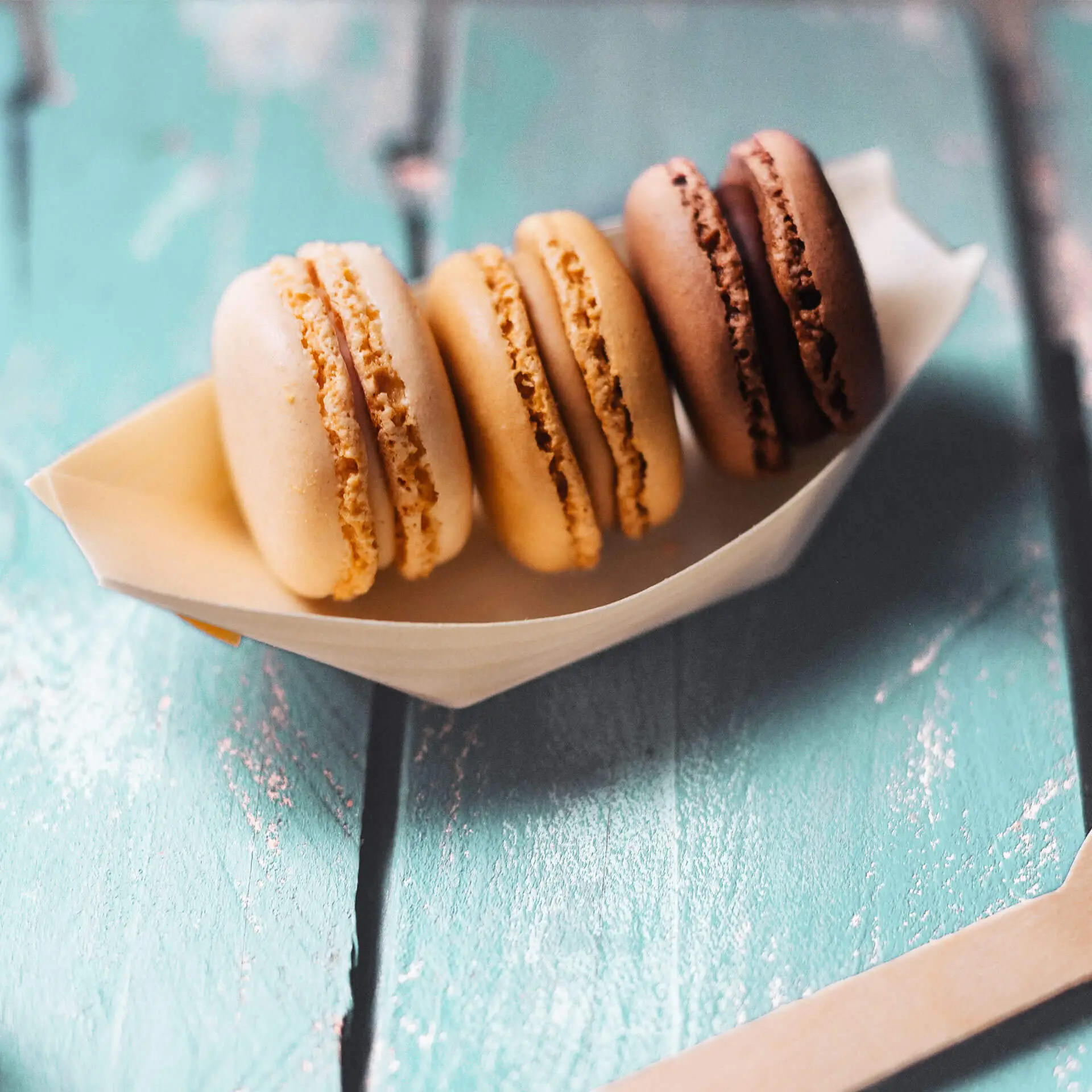 Four macarons in a boat-shaped holder, featuring beige, light brown, dark brown, and chocolate colors on a textured surface.