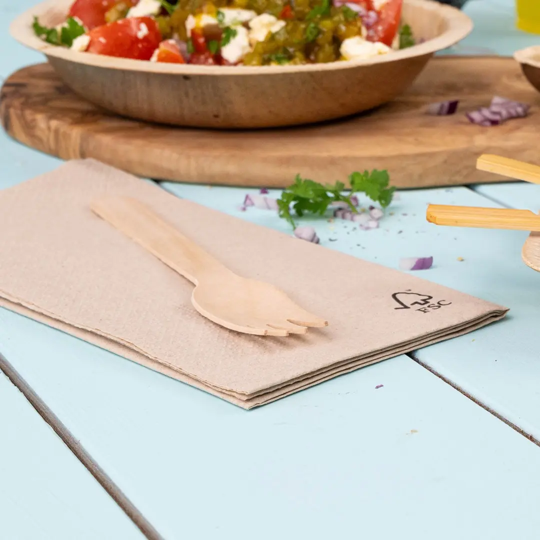 Wooden fork placed on a folded beige napkin, with a bowl of salad in the background on a light blue table.