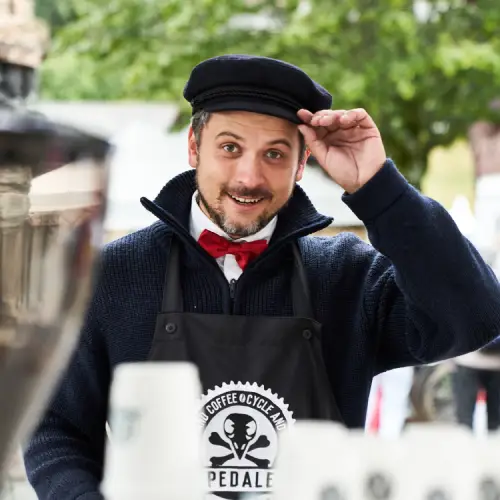 Man in a black cap and bow tie, wearing a branded apron, smiling and greeting visitors at a coffee stand.