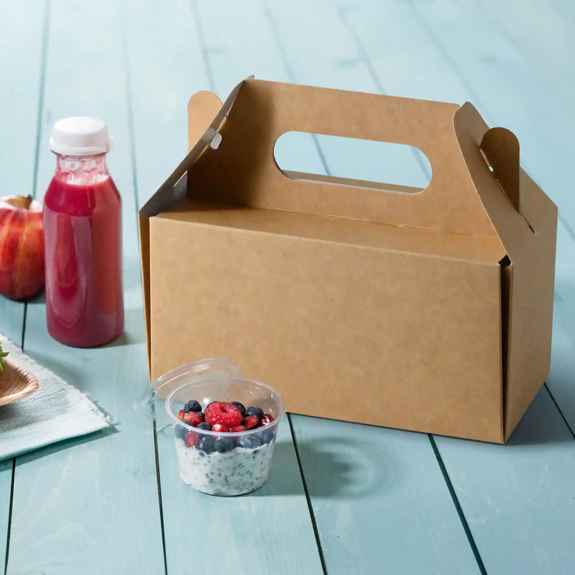 Pappkuchenbox mit Griff, 26 x 17 x 9 cm, neben einem Obstbecher und einer Saftflasche auf einer blauen Holzoberfläche.