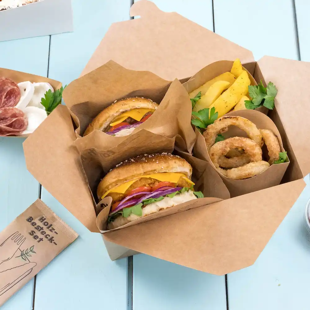 Zwei Hamburger mit Käse, Salat, Tomate und Zwiebel in einer Takeout-Box, begleitet von Zwiebelringen und Pommes.