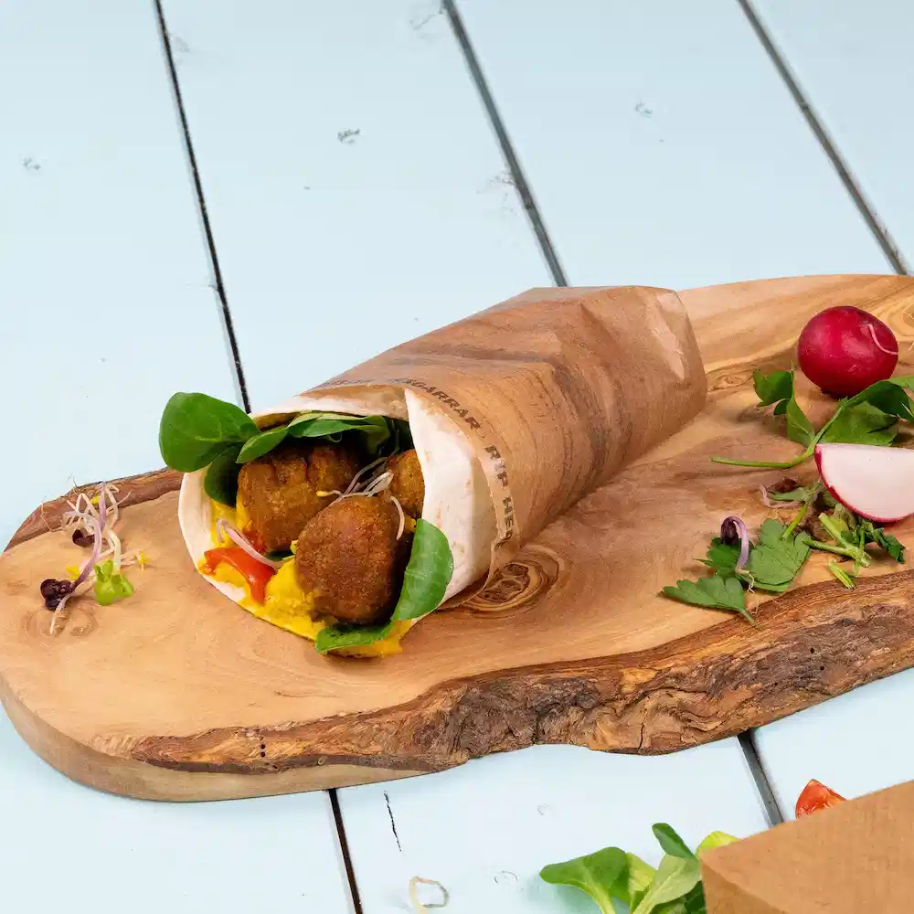 Wrap filled with falafel balls, greens, and sauce on a wooden board, with radish and herbs nearby.