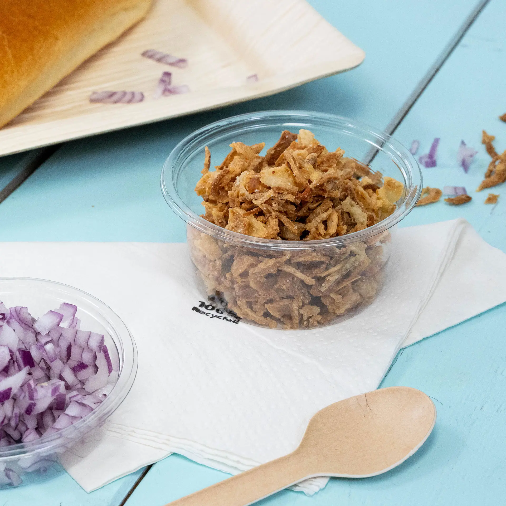 Clear plastic cup with crispy fried onions, bowl of diced red onions, and a wooden spoon on a napkin.