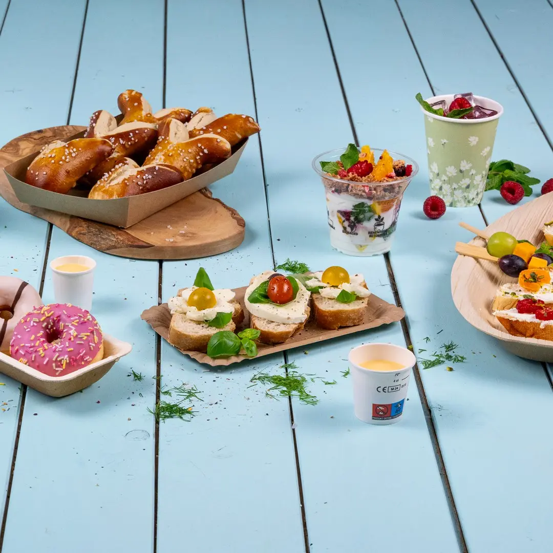 Verschiedene Snacks, darunter Brezeln, belegte Sandwiches, Joghurt-Parfait, Donuts und Obstspieße auf einer blauen Holzoberfläche.