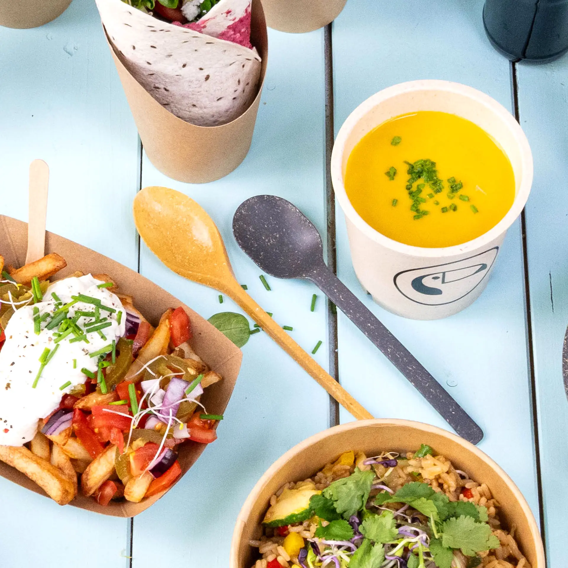 One brown reusable spoon made from bio-compound; bowl of soup, fries with toppings, and a wrapped item displayed nearby.