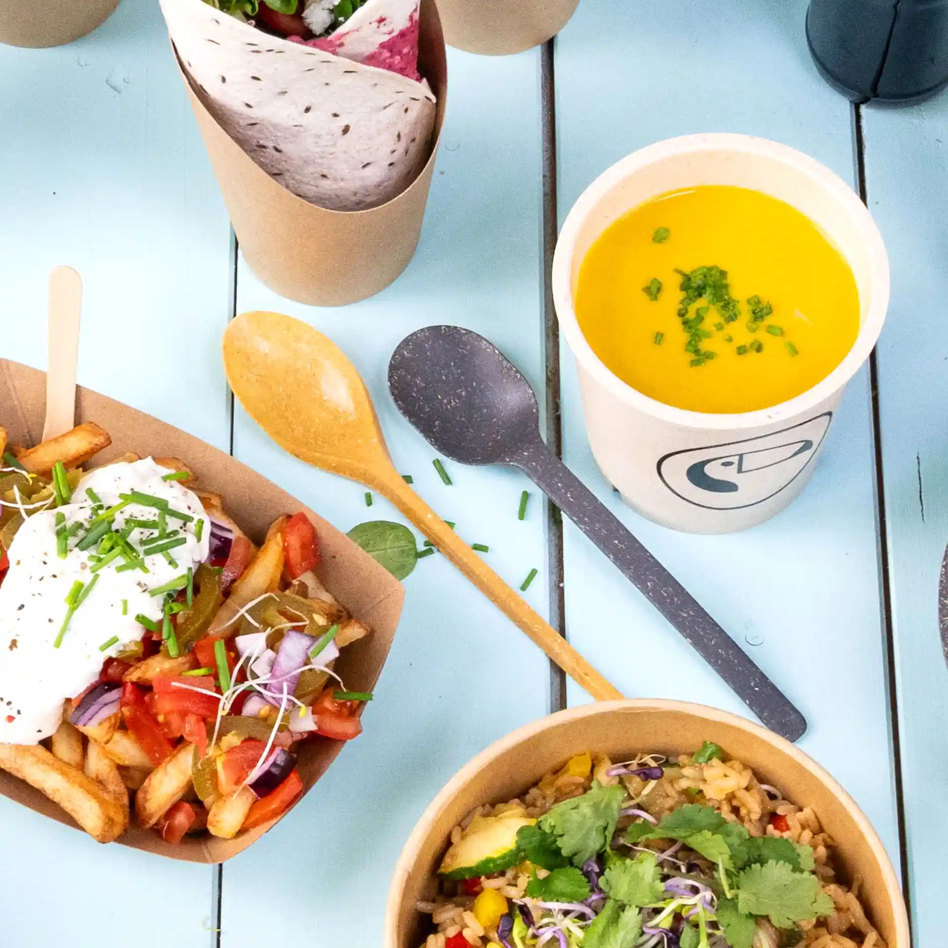 Image of a brown reusable spoon next to food items including fries, soup, and wraps on a blue surface.
