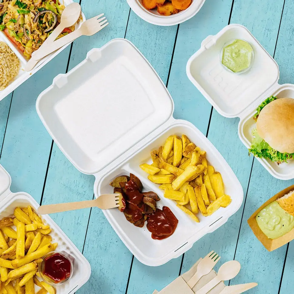 Weiße Takeaway-Behälter mit Pommes, Wurst und Saucen auf einer blauen Holzoberfläche, neben einem Burger und anderen Lebensmitteln.