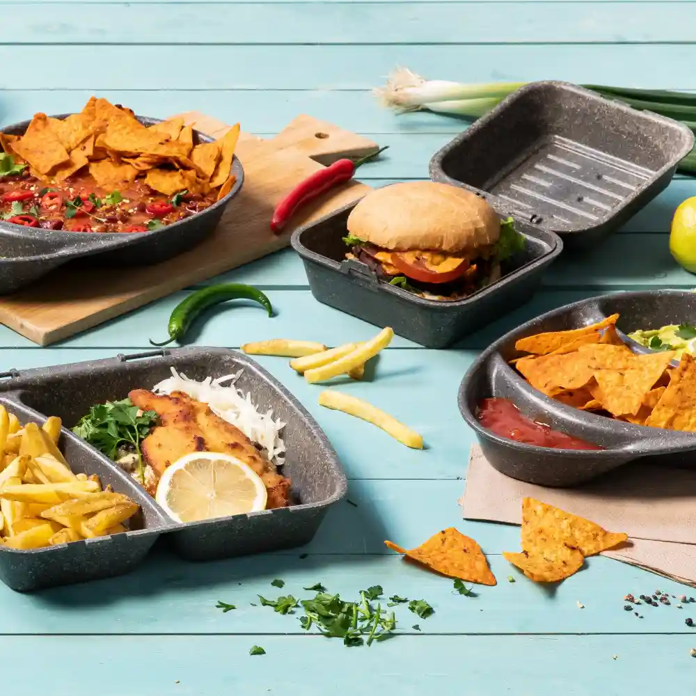 Verschiedene Lebensmittel in wiederverwendbaren Behältern, darunter Nachos, ein Burger, Pommes und Fisch mit Zitrone auf blauer Oberfläche.