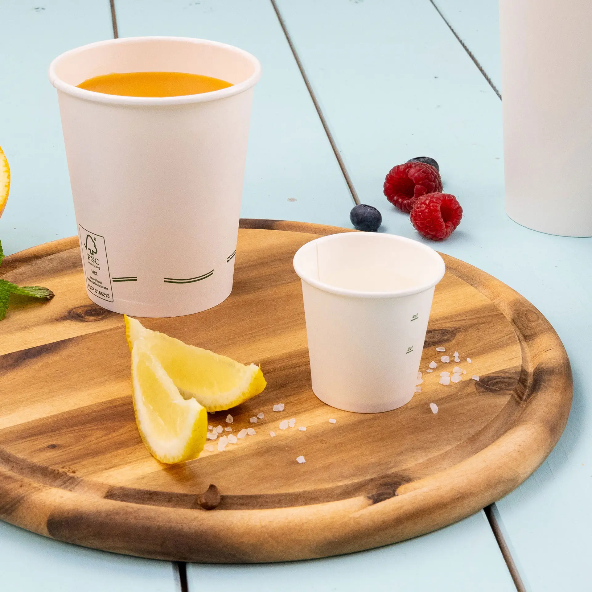 One white 2 oz paper cup on a wooden platter, with a larger cup filled with orange drink, lemon wedges, and berries nearby.