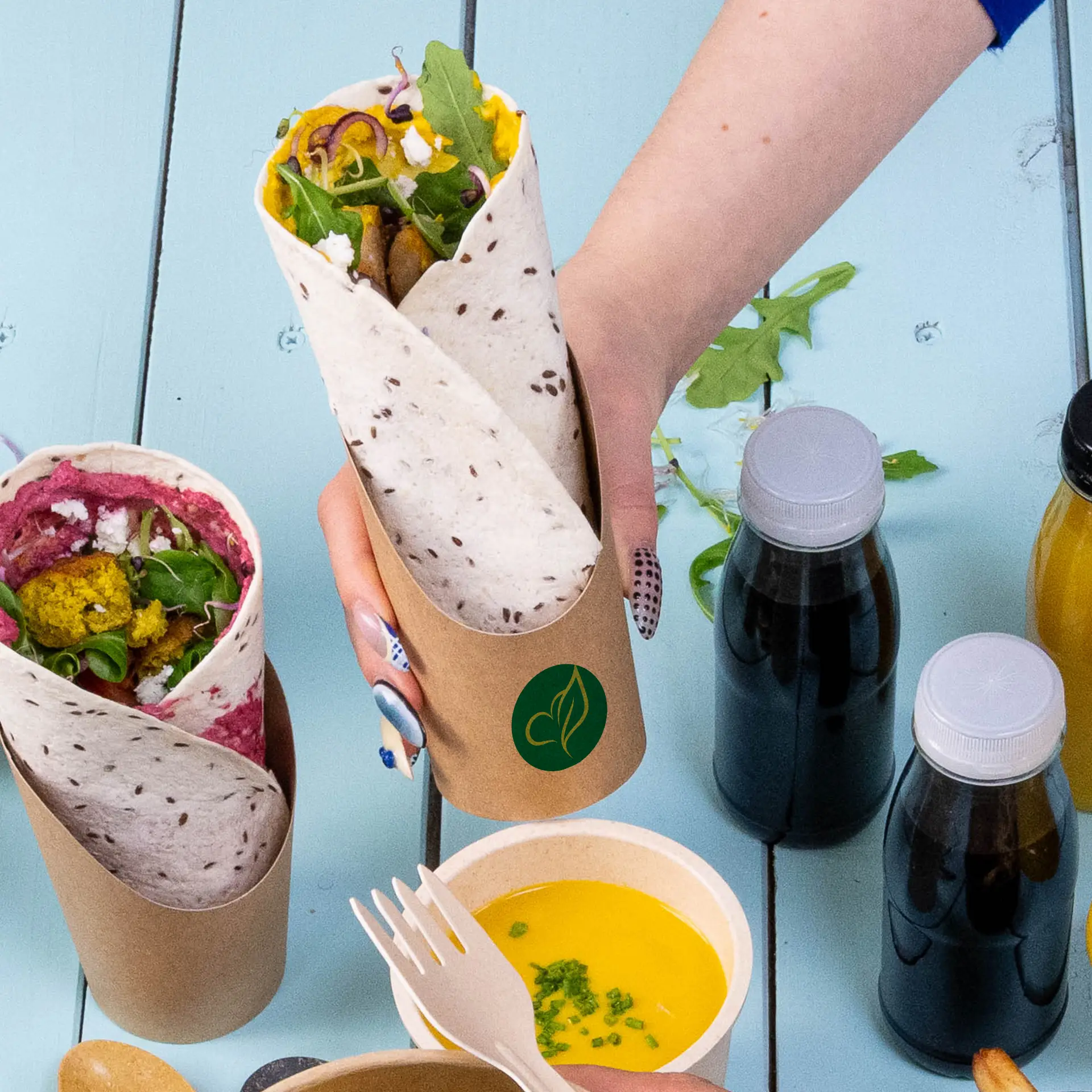 Ein Kraftkarton-Wrapbecher mit gefülltem Wrap; daneben ein Becher mit buntem Wrap, umgeben von Getränken und einer Dipsauce.