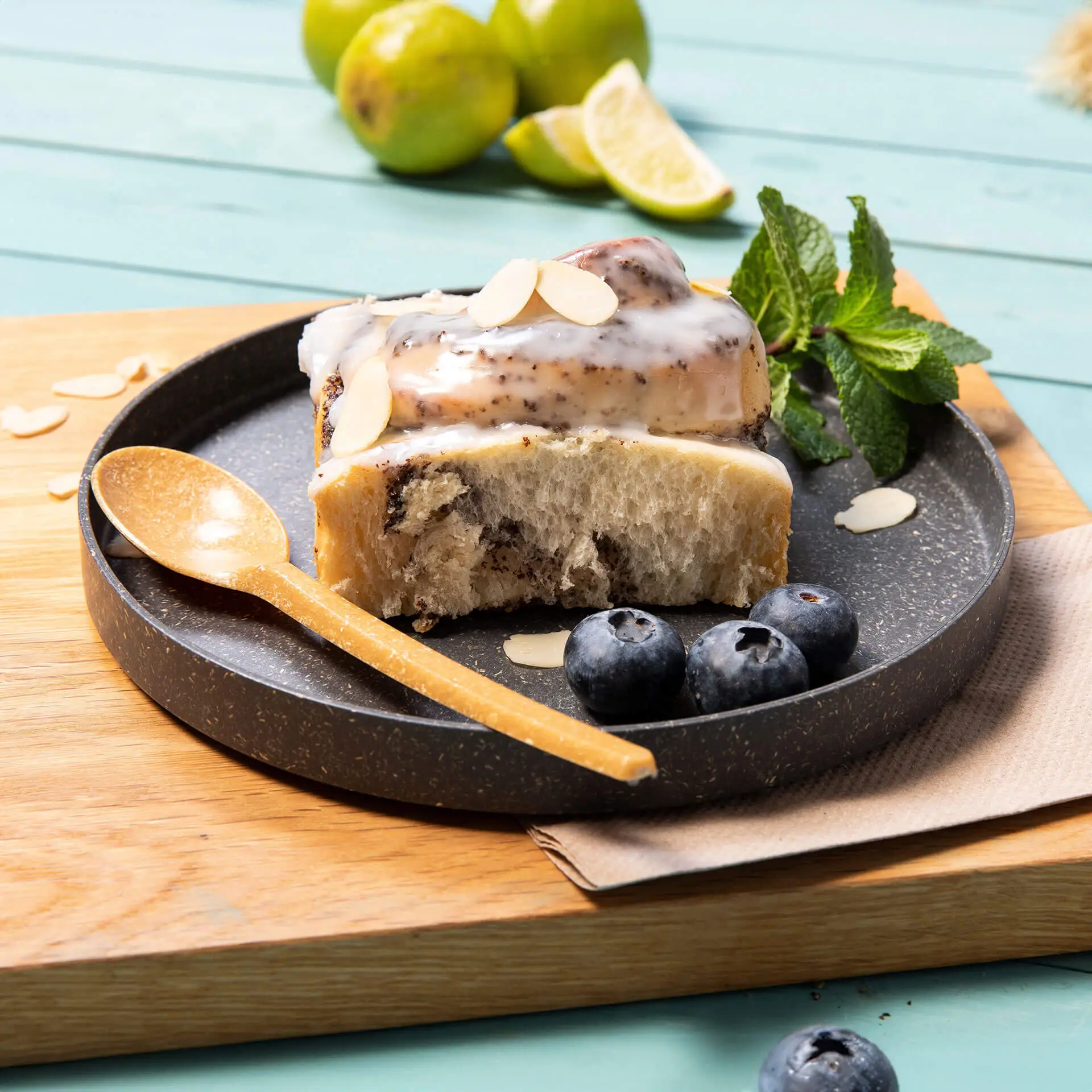Slice of cake topped with icing and almonds on a grey round plate, accompanied by blueberries and a wooden spoon.
