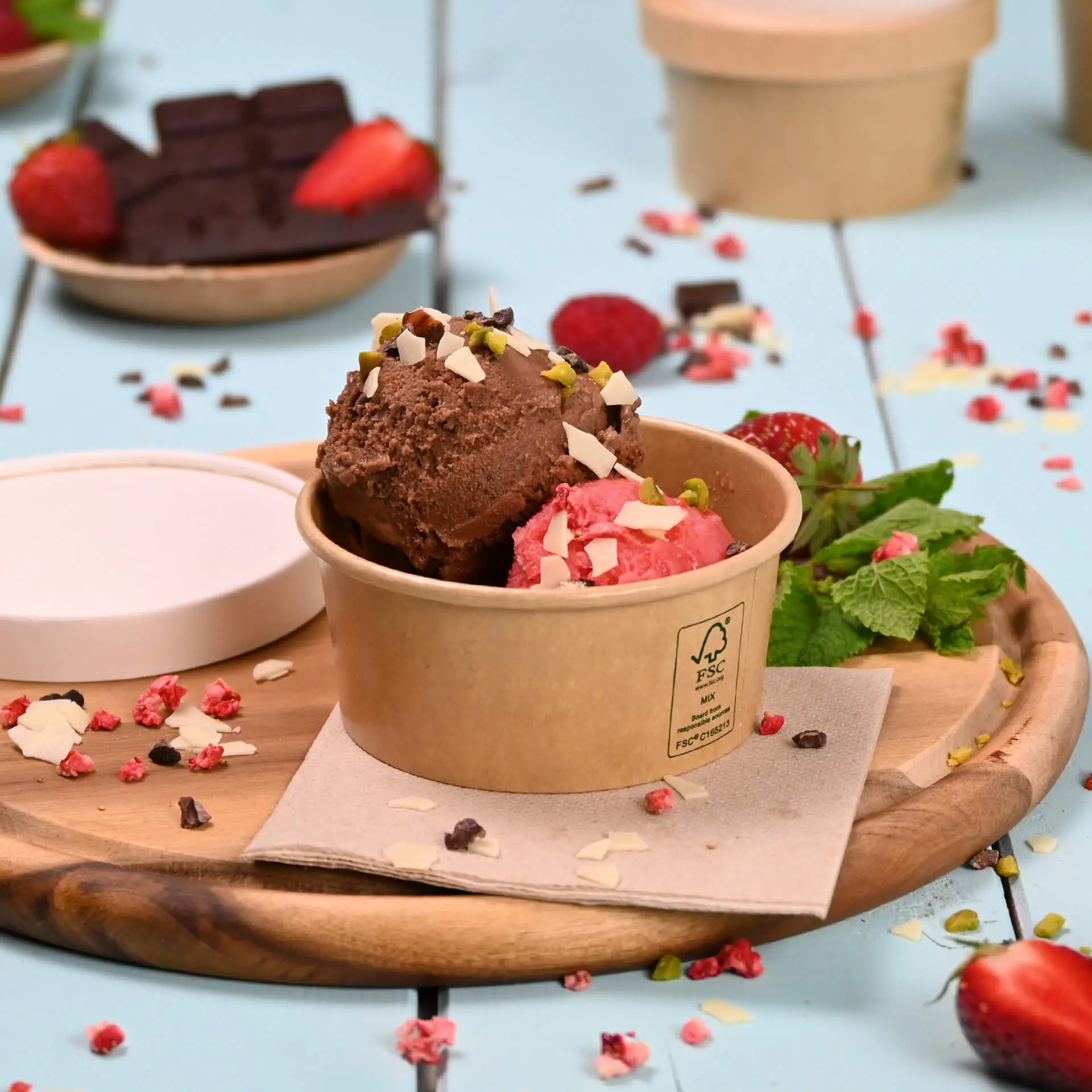 Brauner Papierbecher mit Schokoladen- und Erdbeereis, garniert mit Nüssen, umgeben von frischen Erdbeeren und Minze.