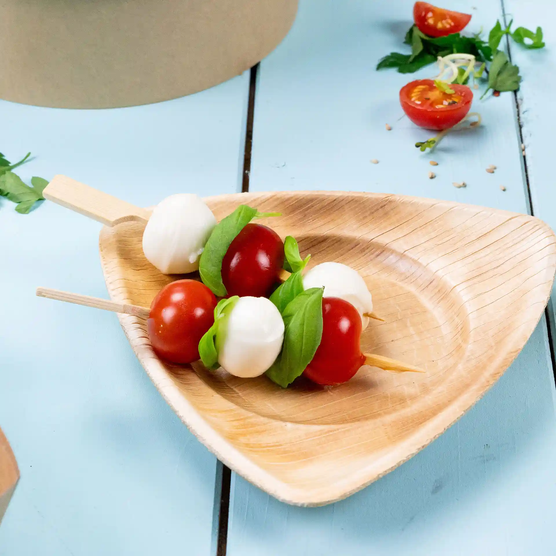 Bambus-Spieße mit Kirschtomaten, Mozzarellakugeln und Basilikum auf einem dreieckigen Holzbrett.