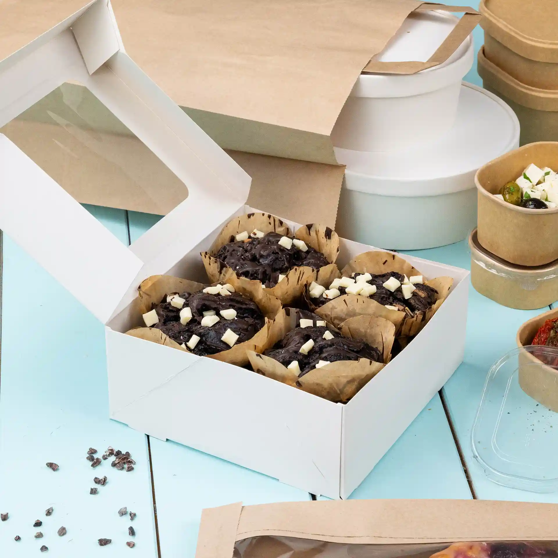 White cardboard box with a clear window containing several chocolate muffins topped with white chocolate pieces.
