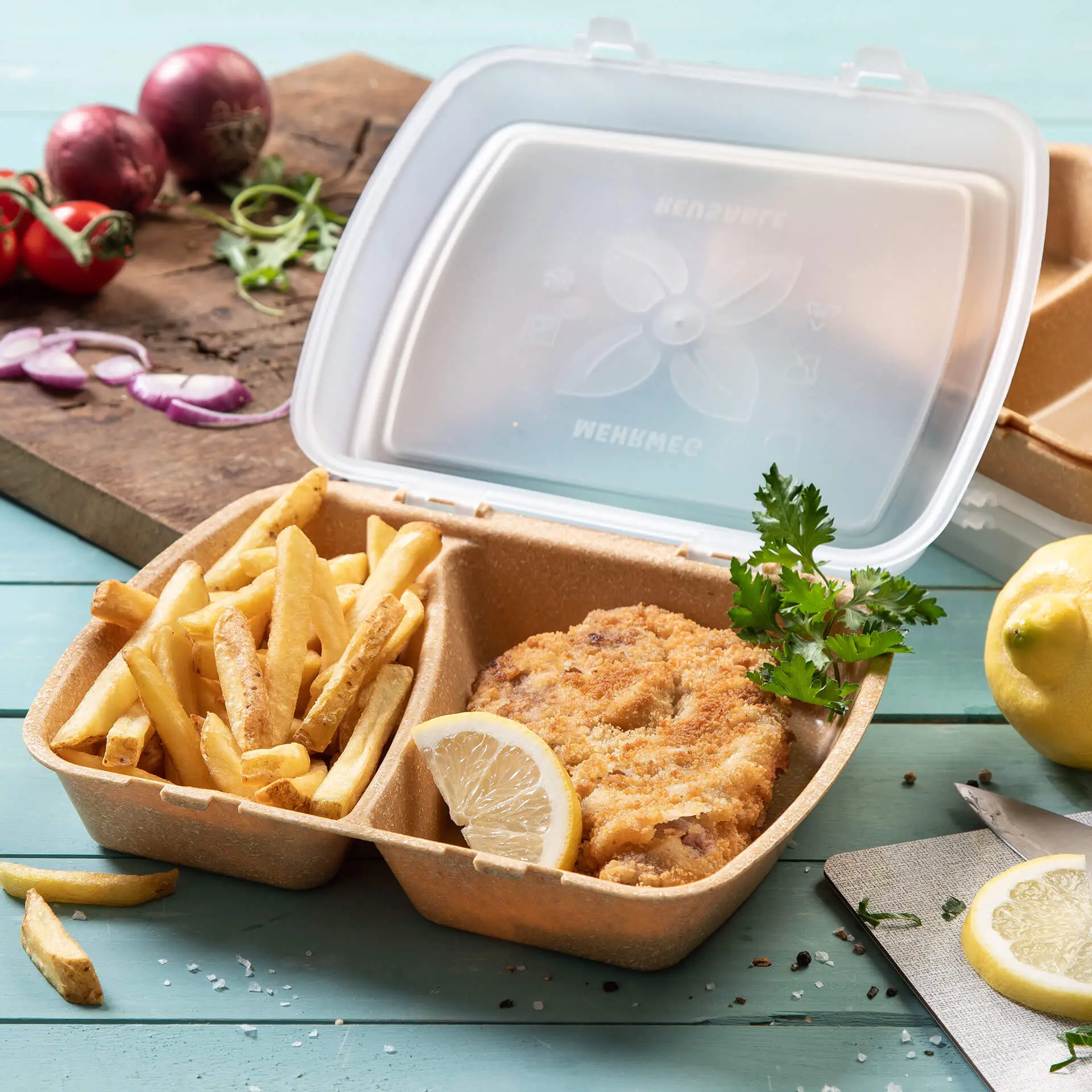Container with fried chicken, fries, and lemon slice, in a transparent plastic box on a blue surface.