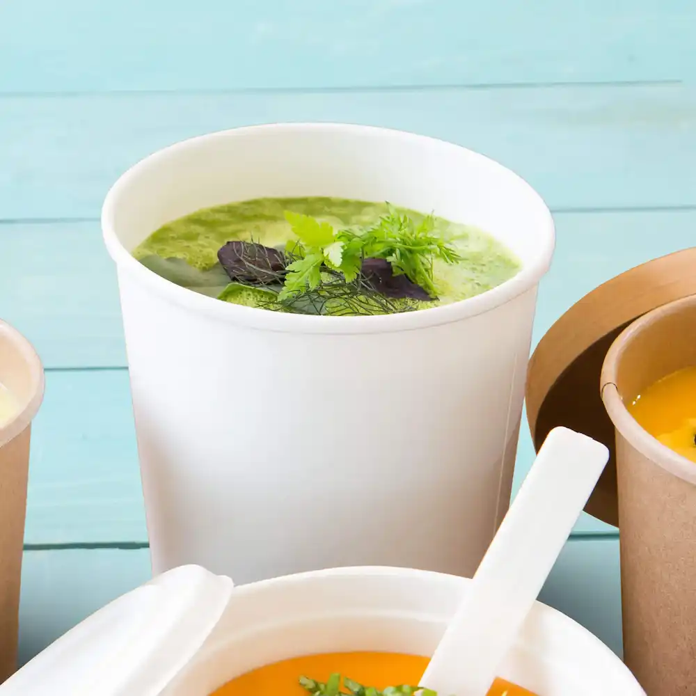 Weißer Suppenbecher, gefüllt mit grüner Suppe und mit Kräutern garniert, umgeben von anderen Behältern.