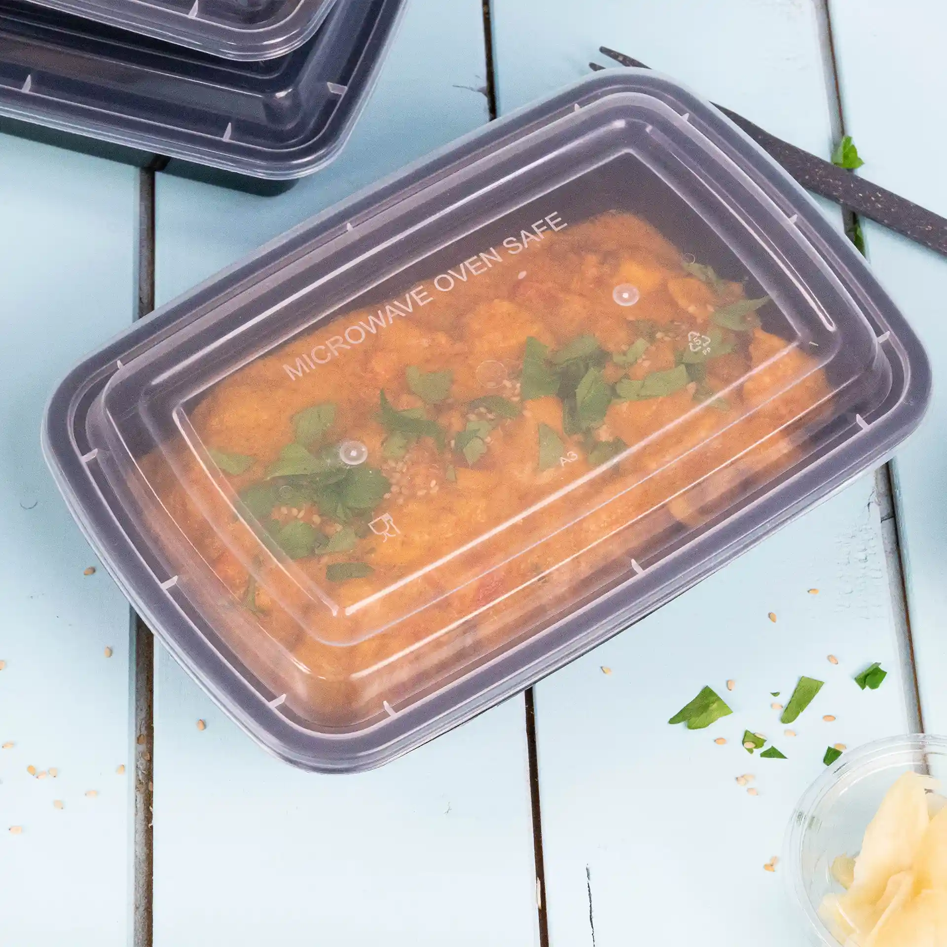 Rechteckiger transparenter Behälter mit einem wiederverwendbaren Deckel, gefüllt mit Essen und mit Kräutern garniert, auf einer blauen Oberfläche.