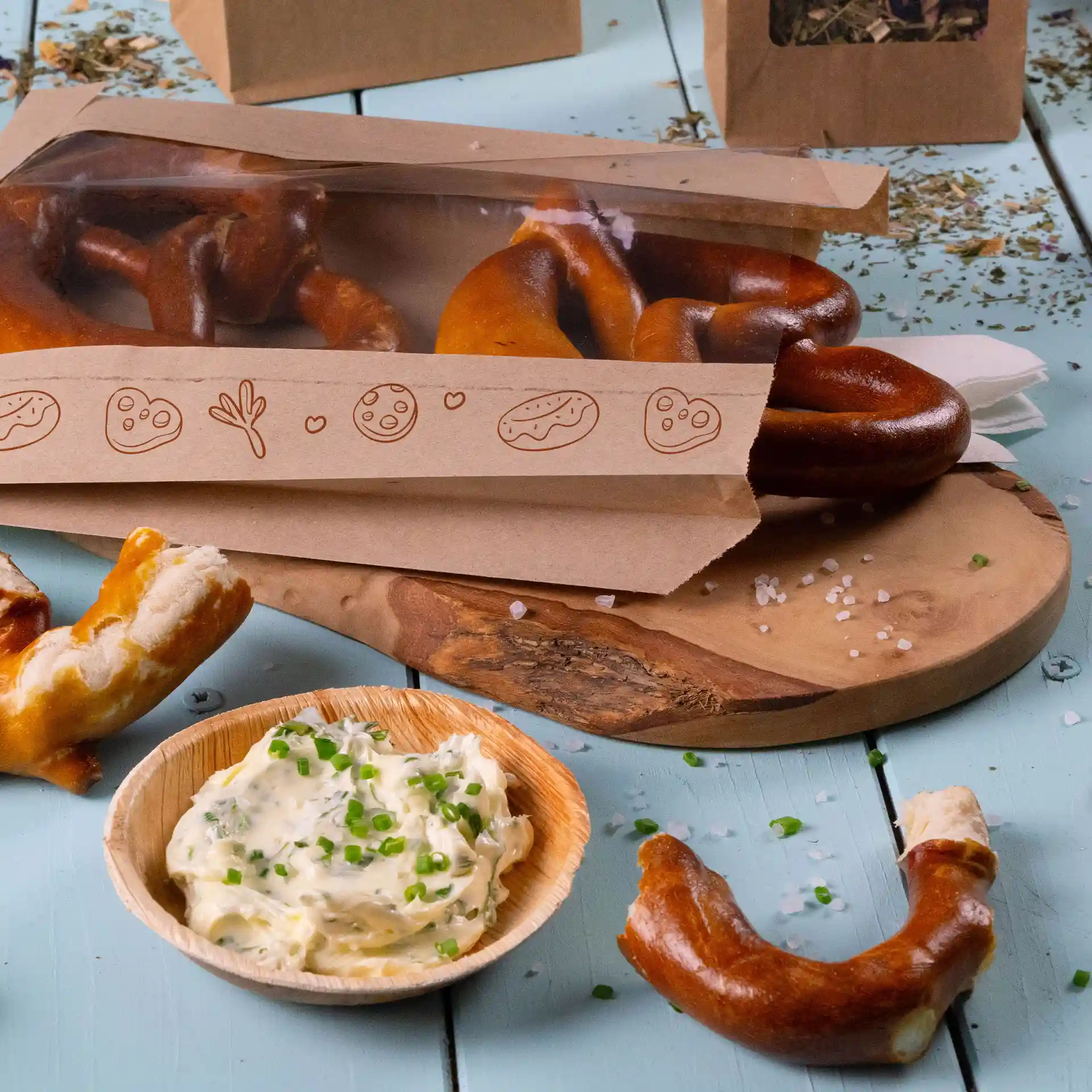 Brown printed paper bag with window containing pretzels, accompanied by a bowl of creamy dip and scattered herbs.