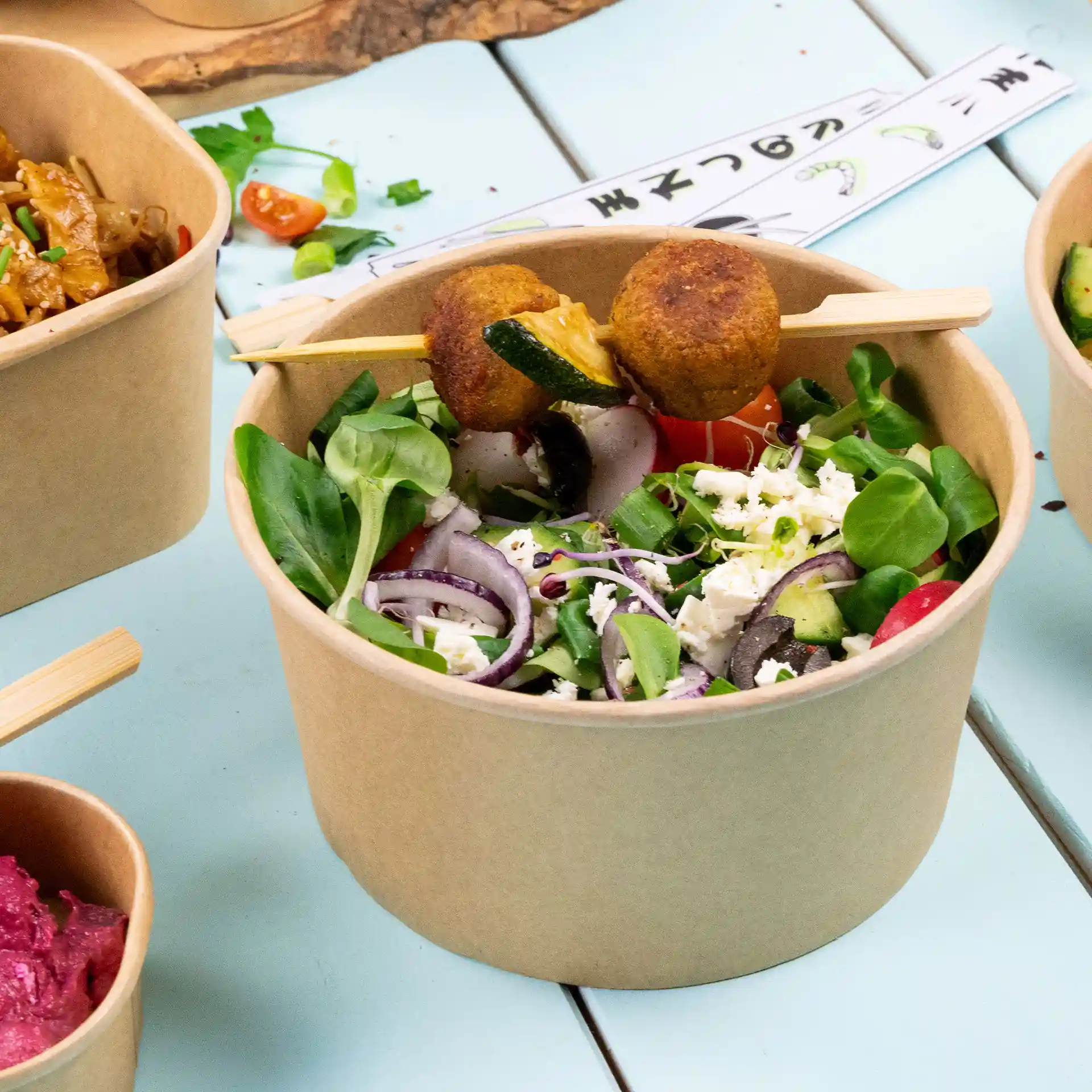 Runde braune Papierschüssel gefüllt mit Salat, garniert mit Spießen von frittierten Speisen, auf einer hellblauen Oberfläche.