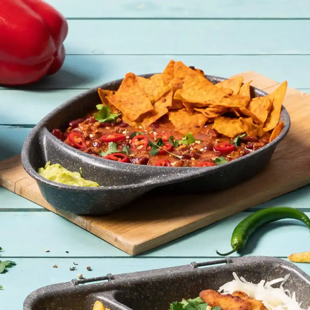 Schüssel mit Chili, belegt mit Tortilla-Chips, geschnittenen roten Chilischoten und Koriander auf einem Holzbrett mit Belag.