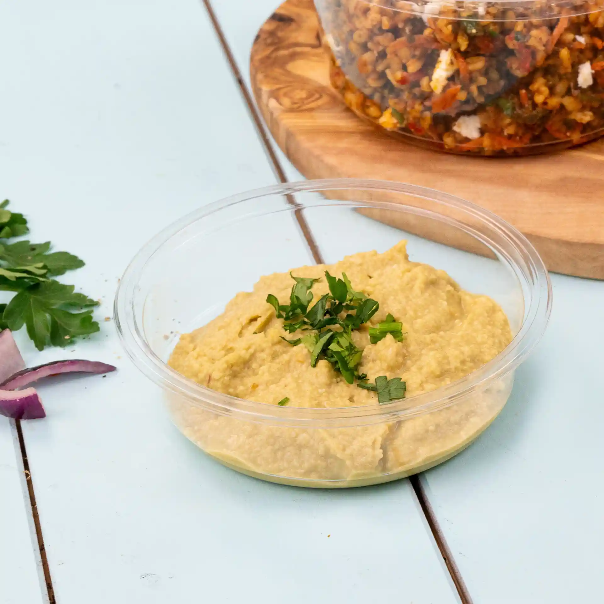 Round transparent deli container with hummus topped with chopped parsley, placed on a light blue surface.