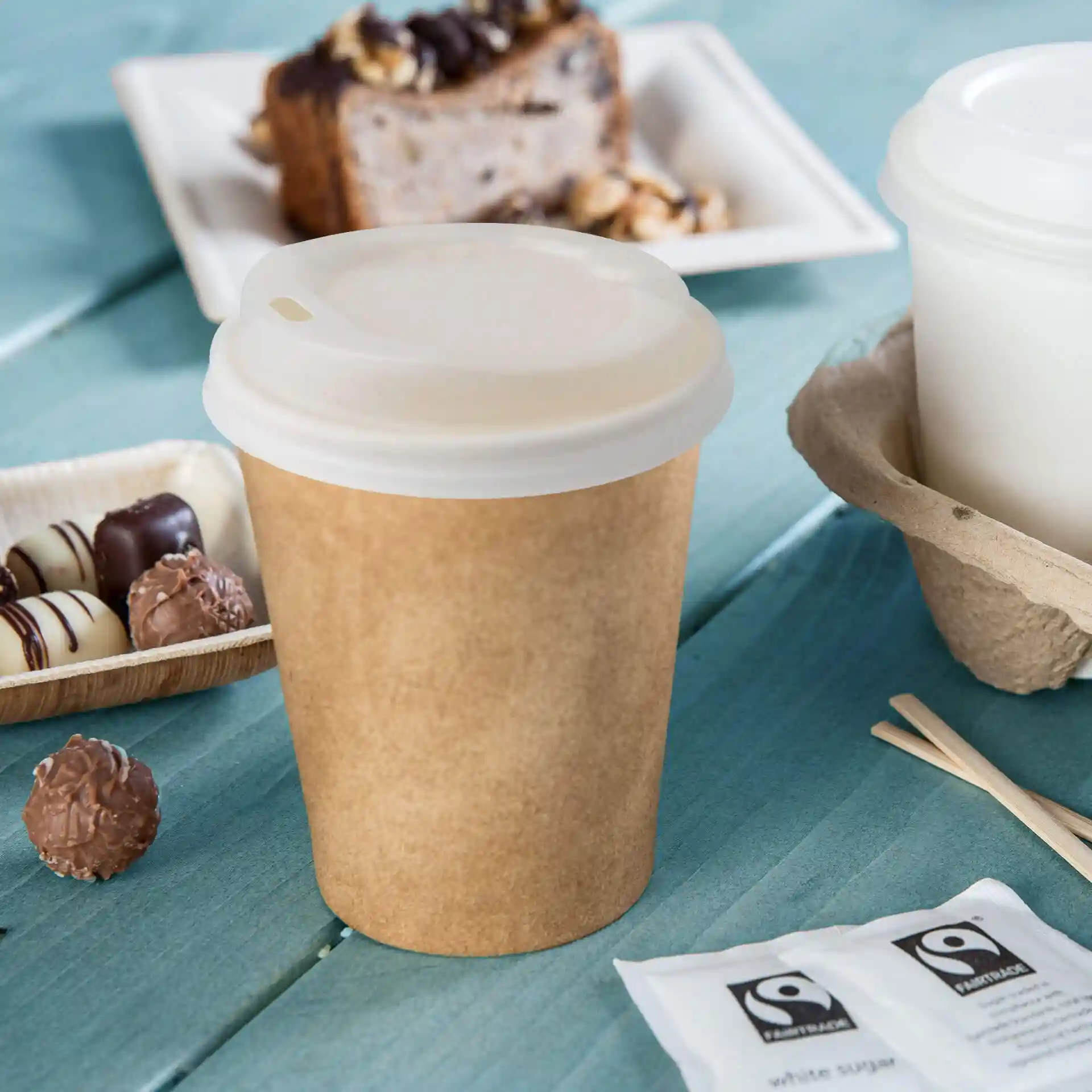 Brown paper cup with a white lid, placed on a blue wooden surface, surrounded by chocolates and sugar packets.