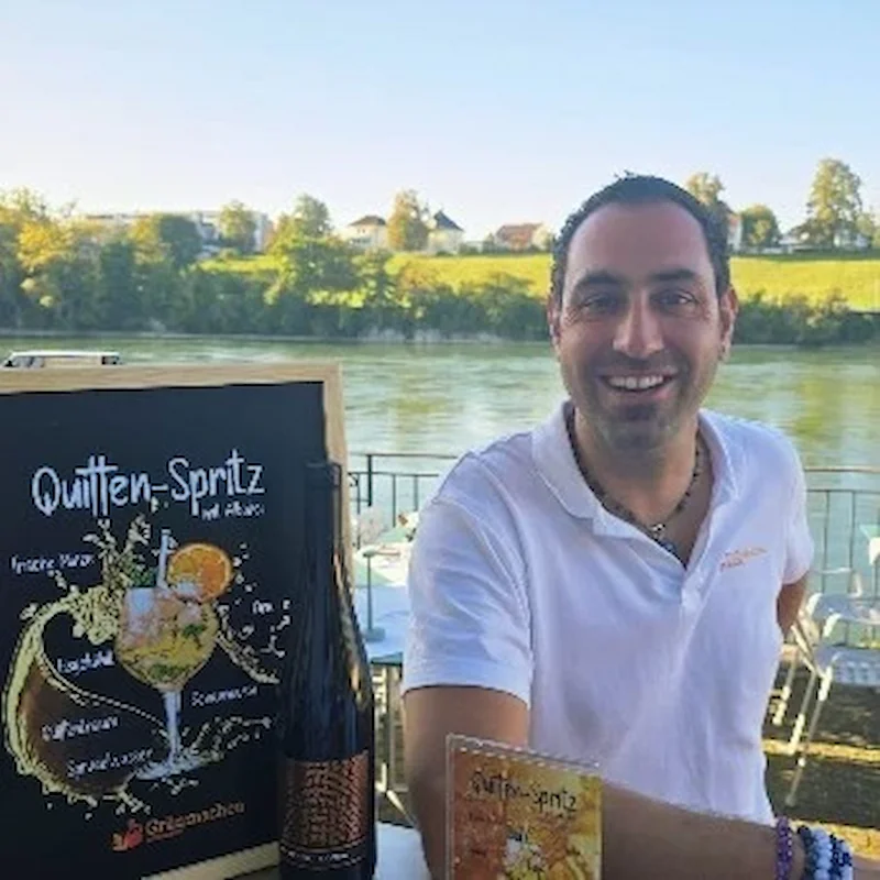 Man smiling behind a bar with a sign for "Quitten-Spritz," a drink, and a bottle of beverage on display by a river.