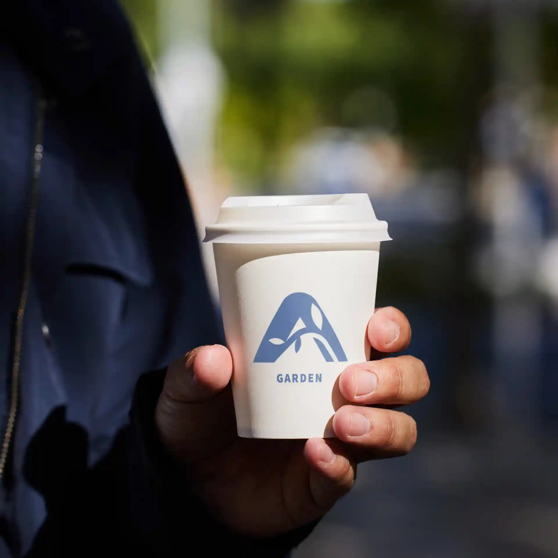 White paper cup with a blue logo and the word "GARDEN," held in a hand, outdoors with a blurred background.