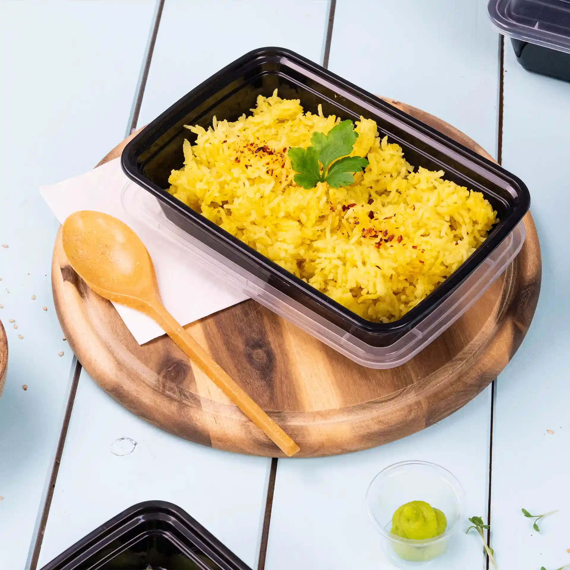 Container with yellow rice topped with a green herb, wooden spoon beside it, on a round wooden board.