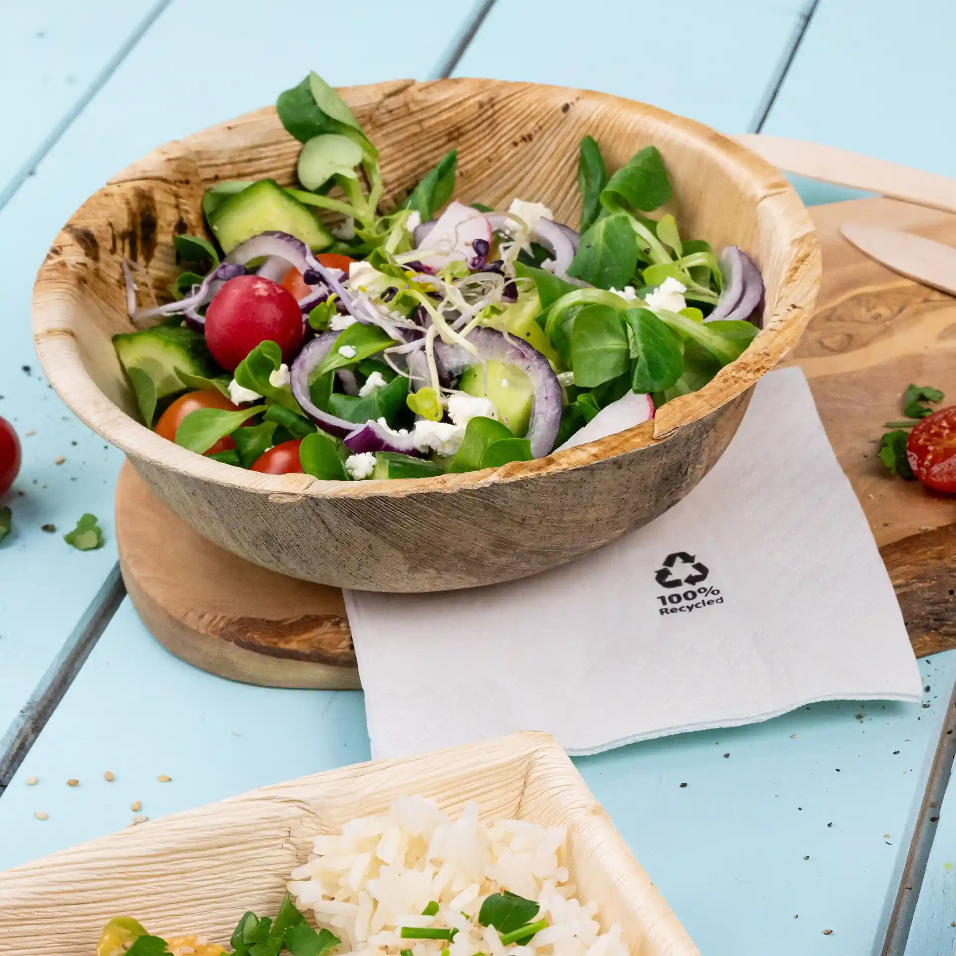 Runde Palmblatt-Schüssel gefüllt mit gemischtem Salat, auf einem Holzbrett neben einer Serviette und Reis in einem Behälter.