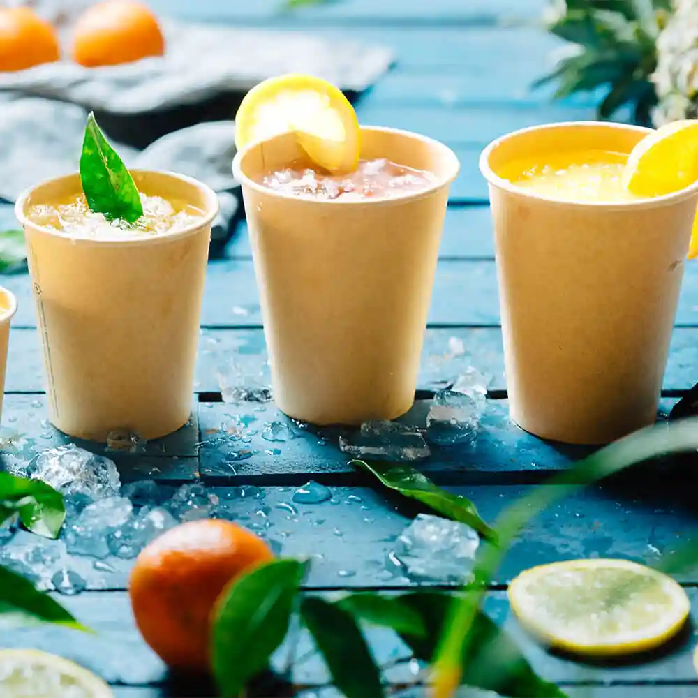Drei braune Pappbecher (12 oz) mit Getränken, garniert mit Zitrone und Blättern, auf einer blauen Holzoberfläche mit Eis.