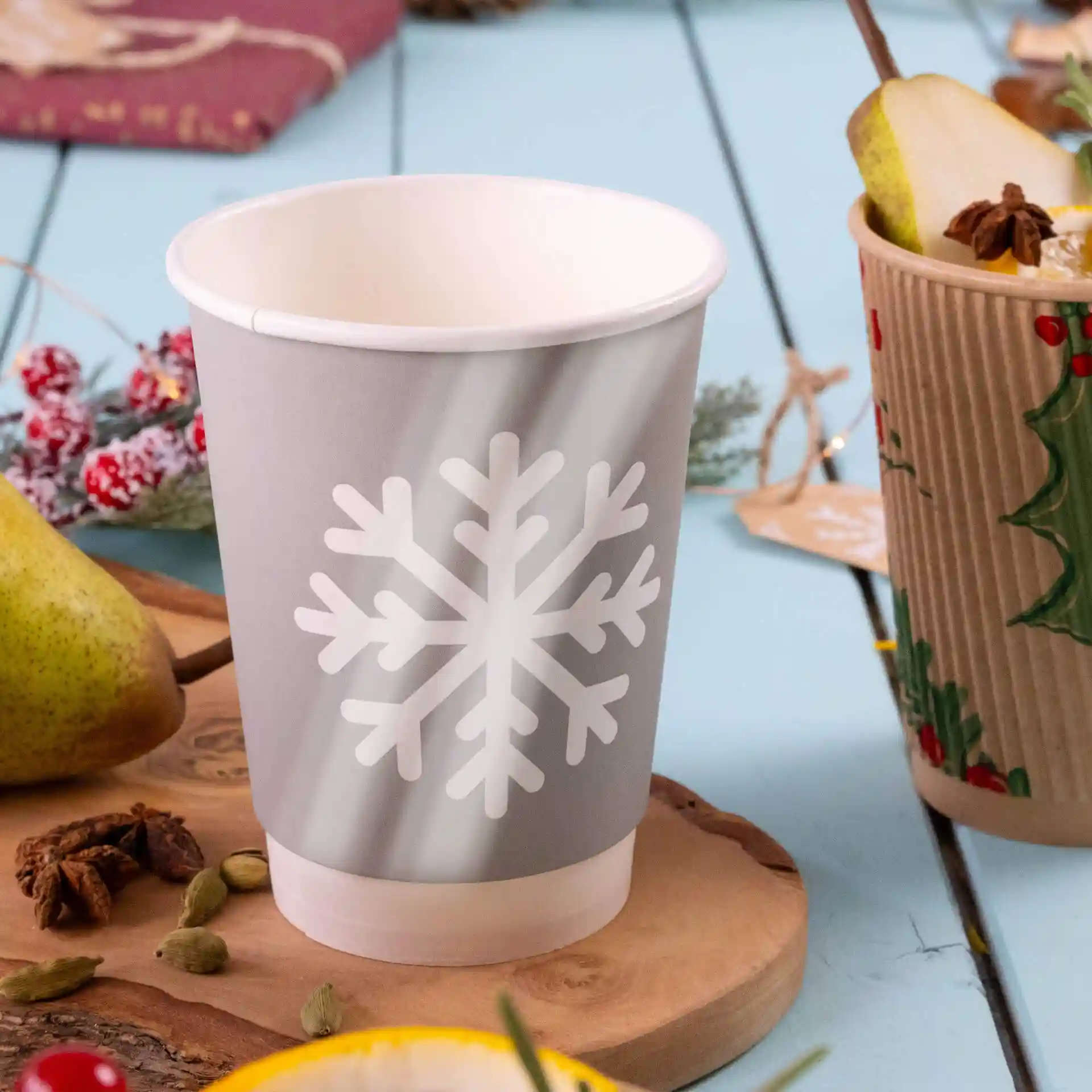 Grauer Becher mit weißem Schneeflocken-Design, doppelwandig, auf einer Holzoberfläche mit einer Birne und Gewürzen.