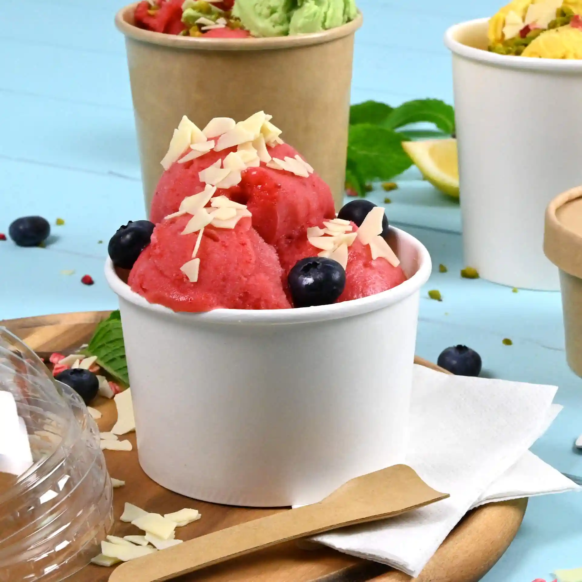 Weißer Becher mit rosa Eiscreme, garniert mit Mandelstückchen und Heidelbeeren, auf einer Holzoberfläche.
