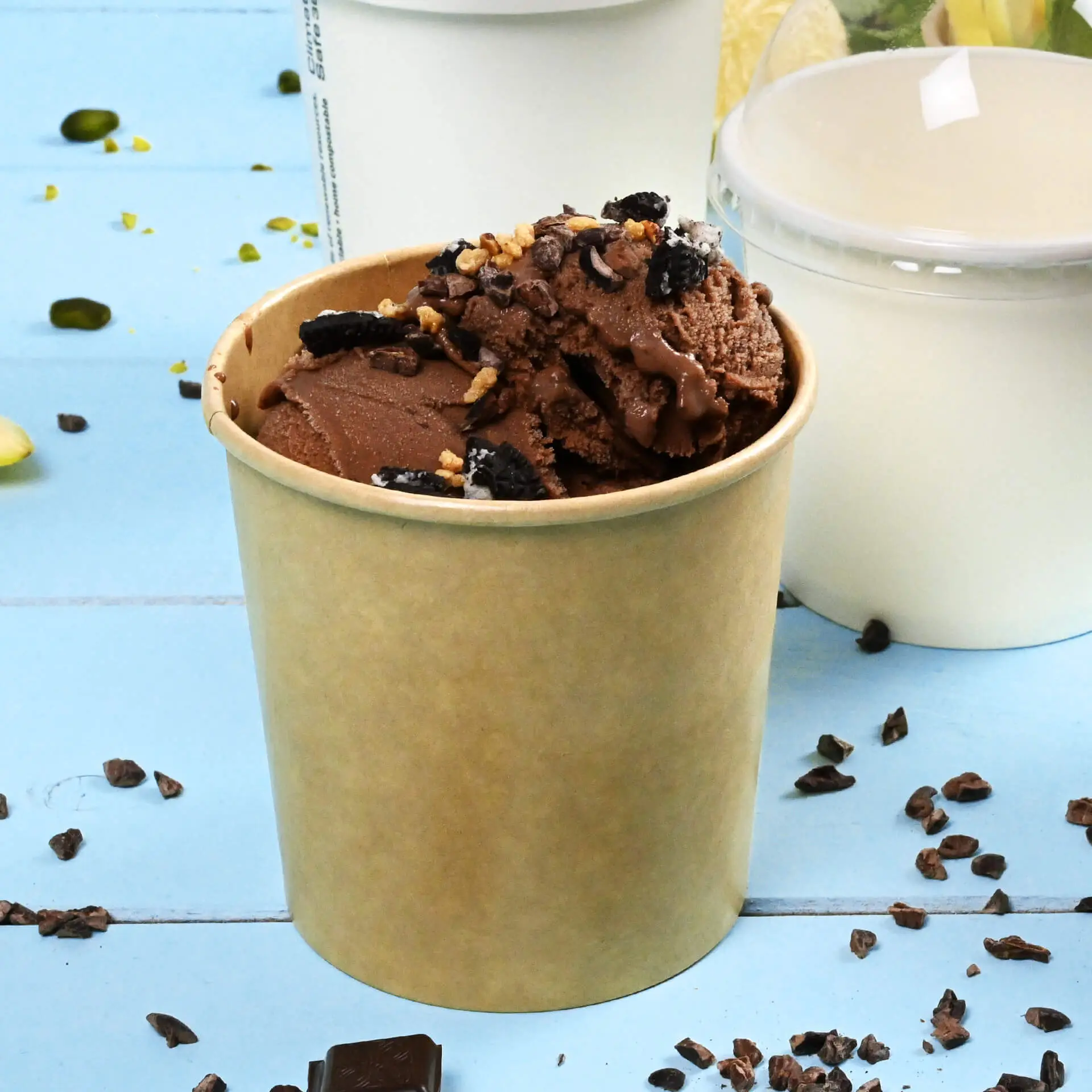 One brown paper ice cream cup filled with chocolate ice cream and toppings, placed on a blue surface with scattered toppings around.