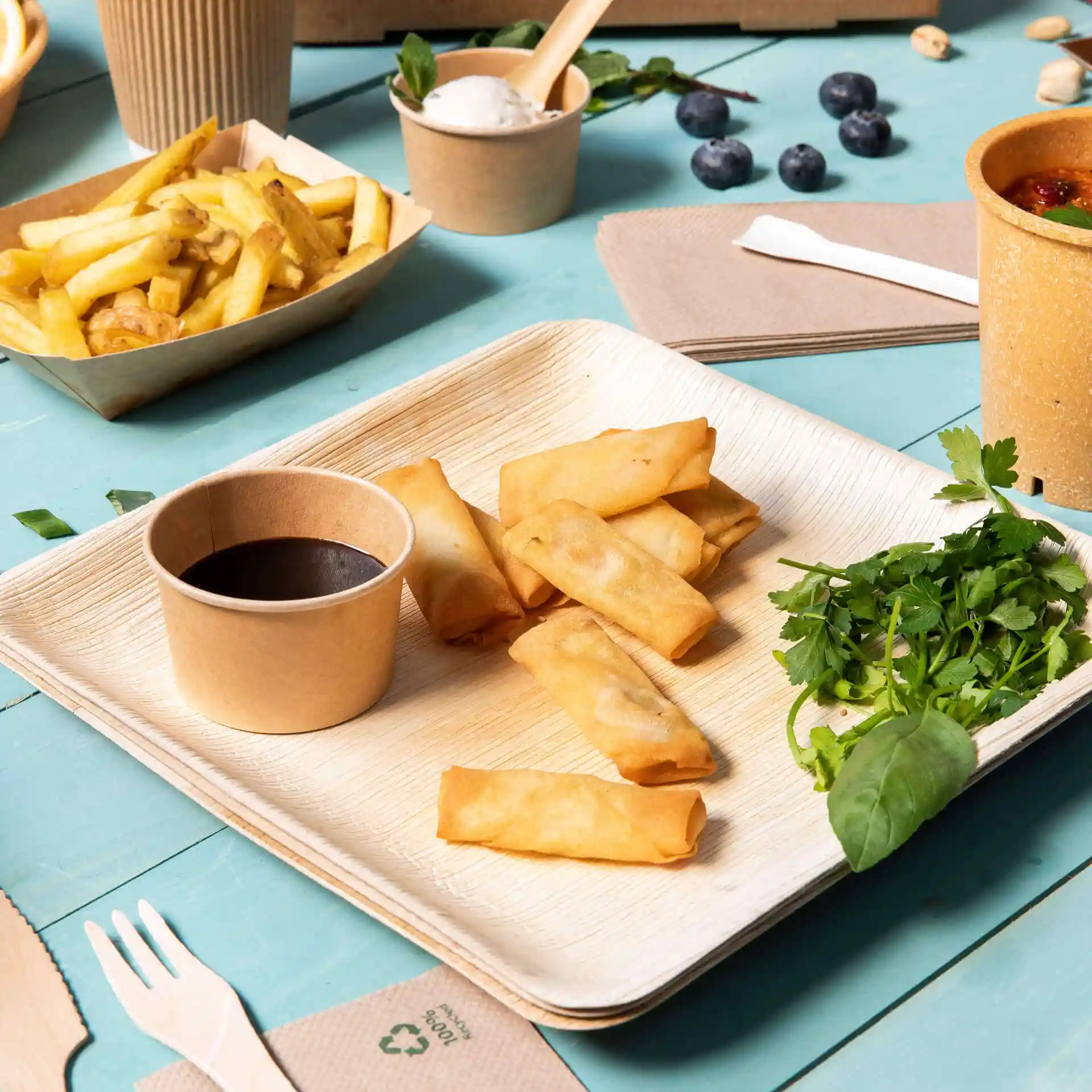 Ein quadratischer Palmblatt-Teller mit Frühlingsrollen, einer Schale Sauce, frischen Kräutern und einer Portion Pommes auf einem blauen Tisch.