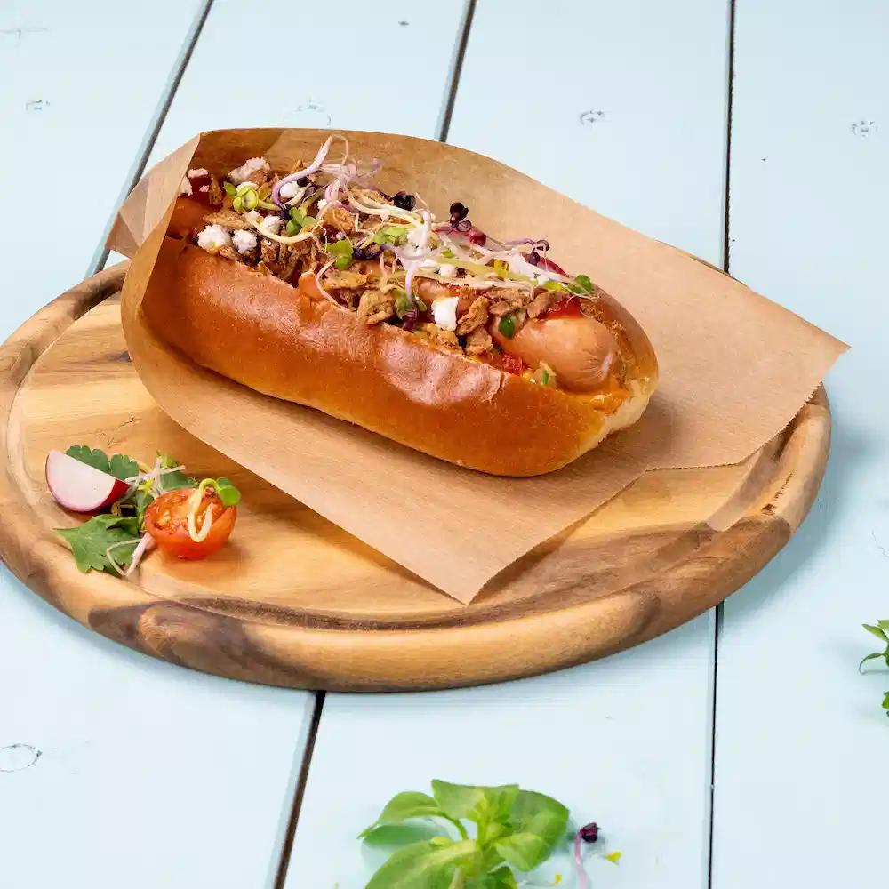 Hotdog im Brötchen mit Zwiebeln, Kräutern und knusprigen Toppings auf einem Holztablett mit Beilagen.
