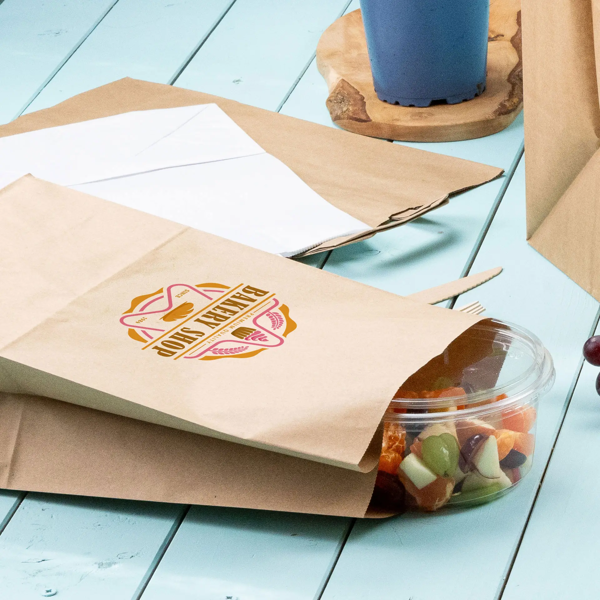 Braune Papiertüten mit einem kreisförmigen Design und einem klaren Plastikbehälter mit gemischtem Obst auf einer blauen Oberfläche.